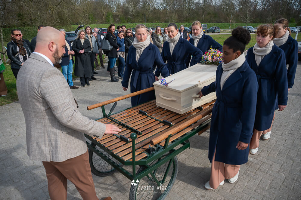 Vrouwelijke dragers begeleiden de uitvaartkist naar de baarwagen tijdens een persoonlijke uitvaart, vastgelegd door een uitvaartfotograaf.